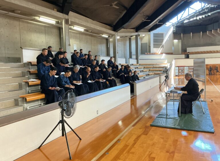 観覧席で資料を手に講師の話を聞く受講者